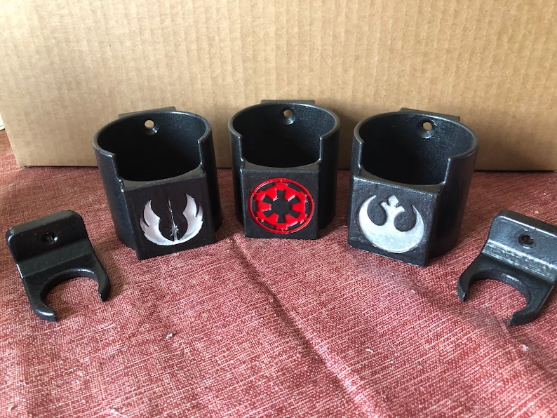 May include: Three black wall-mounted cup holders with different Star Wars logos. The logos are the Rebel Alliance symbol, the Imperial symbol, and the symbol of the Mandalorian faction. Each holder has a small hook on the side.