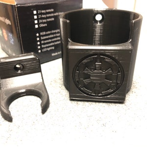 May include: Black plastic wall mount with a hook for hanging a mug or other object. The mount has a small hole for attaching to a wall.  The second image shows a black plastic cup holder with a Star Wars Imperial symbol on the front. The cup holder has a hole at the top for hanging.