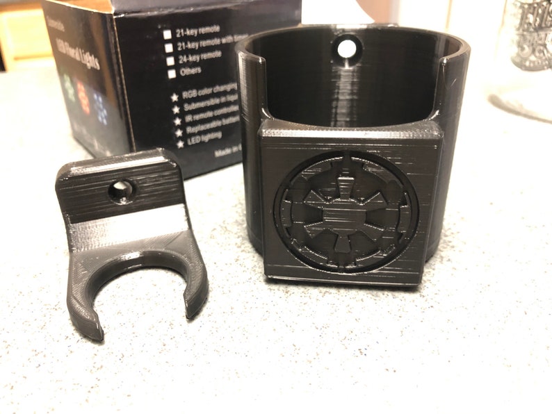 May include: Black plastic wall mount with a hook for hanging a mug or other object. The mount has a small hole for attaching to a wall.  The second image shows a black plastic cup holder with a Star Wars Imperial symbol on the front. The cup holder has a hole at the top for hanging.