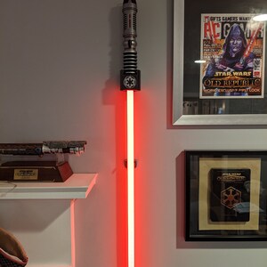 May include: A red lightsaber with a black and silver hilt is mounted on a wall. The lightsaber is lit and the blade is red. The lightsaber is mounted on a wall with a black and silver mount. The lightsaber is a Star Wars themed item.