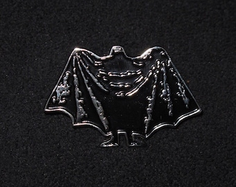 Puede incluir: Un pin de metal plateado con forma de murciélago con una silueta negra. El murciélago tiene una capa y está de pie con las alas extendidas.