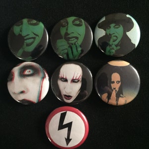 May include: Set of eight round pinback buttons featuring images of Marilyn Manson. The buttons are black with white, red, and green accents. One button features a black and white symbol of a lightning bolt in a circle.