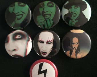 May include: Set of eight round pinback buttons featuring images of Marilyn Manson. The buttons are black with white, red, and green accents. One button features a black and white symbol of a lightning bolt in a circle.