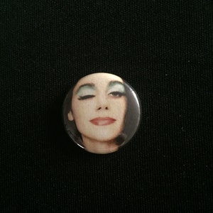 May include: A round button featuring a close-up image of a woman's face. She has dark hair and is wearing green eyeshadow. Her mouth is slightly open and she is looking at the camera.