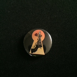 May include: A black and gold button with a keyhole design. A gun with a rose growing out of the barrel is inside the keyhole. The background is a sunset orange.