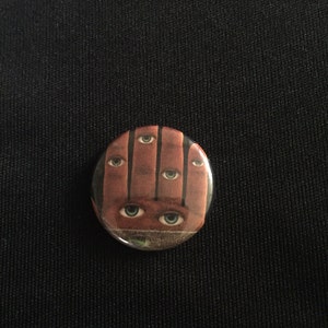 May include: A round button with a brown hand with five eyes on it. The eyes are green and the hand is brown.
