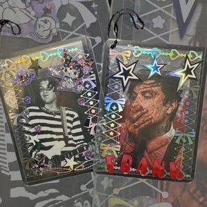 Könnte beinhalten: Zwei rechteckige Fotohalter aus klarem Kunststoff mit schwarzen Kugelkettchen. Der linke Halter zeigt eine Person im gestreiften Hemd und einen lila Aufkleber. Der rechte Halter zeigt eine Person mit roten Markierungen und dem roten Schriftzug FRANK.
