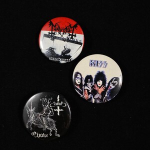 May include: Three round black and white button pins. The first pin features a red, white, and black logo with the text "Mayhem" and "Deathcrush". The second pin features a black and white photo of four people with the text "KISS". The third pin features a black and white illustration of a creature with horns and the text "Evoke" and "Robbot".