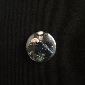 May include: A round button featuring a black and white image of a man with long black hair, wearing a black leather jacket and a black shirt. The man is holding a small object in his hands.