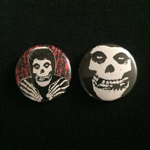 以下が含まれることがあります： 2つの丸いボタン。片方は赤と黒の背景に白黒の骸骨のイラスト、もう片方は黒い背景に白いドクロのイラスト。どちらのボタンも光沢のある仕上がりです。