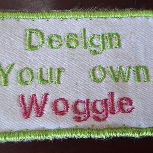 May include: A white fabric patch with green edging and the embroidered text "Design Your own Woggle" in green and pink. Two red buttons are attached to the patch.