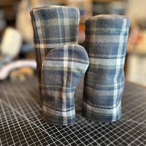 May include: Three plaid fabric items in shades of blue, gray, and white. The items appear to be cylindrical, possibly covers or sleeves. The fabric has a classic plaid pattern. The items are arranged on a black surface with a grid pattern.