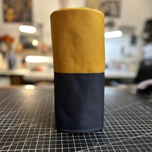 May include: A cylindrical container, divided into two sections. The top half is mustard yellow, and the bottom half is navy blue. The container is made of a sturdy fabric, and it stands upright on a black surface with white grid lines.