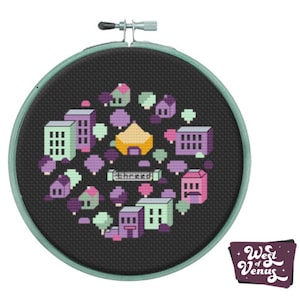 May include: A black embroidery hoop with a colorful pixelated design of a city with buildings, trees, and a yellow structure. The text "Shrecko" is visible in the design. The hoop is surrounded by a light green border.