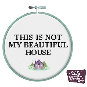 May include: A white embroidery hoop with the text "This is not my beautiful house" and a pixelated image of a house in the centre. The hoop is green and has a metal clamp.