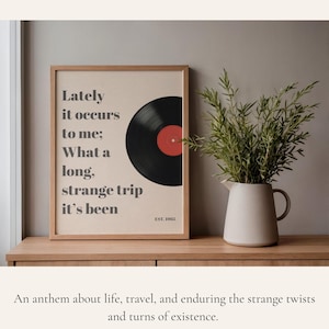 May include: Framed print featuring a black vinyl record with a red center. The print has the text "Lately it occurs to me; What a long, strange trip it's been" and "EST. 1965". A white pitcher with green foliage sits to the right.