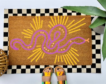 May include: A rectangular brown coir doormat featuring a central purple snake illustration with orange spots, surrounded by yellow sunburst rays. The mat is layered over a black and white checkered rug.