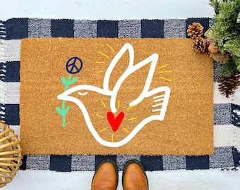 May include: A brown coir doormat with a white outline of a dove holding a green flower with a peace sign. The dove has a red heart in its chest and yellow rays of light around it.