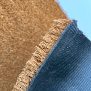 May include: Close-up of a brown coir doormat with a black rubber backing. The coir fibers are frayed at the edge of the mat.