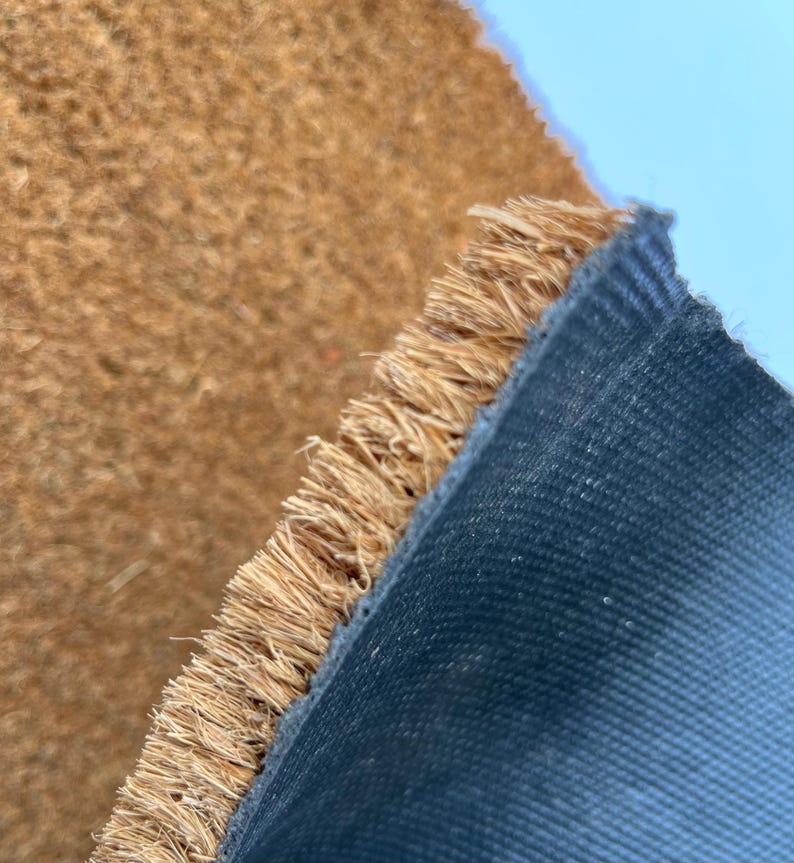 May include: Close-up of a brown coir doormat with a black rubber backing. The coir fibers are frayed at the edge of the mat.