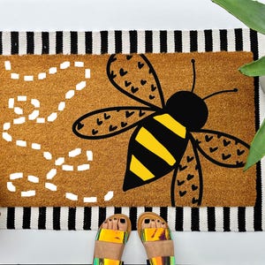 May include: A rectangular doormat with a large bee illustration. The bee has yellow and black stripes and black heart-shaped details on its wings. The mat has a brown center and a black and white striped border.