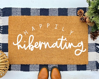 May include: A brown coir doormat with the text "Happily Hibernating" in white cursive lettering. The doormat is on a black and white checkered rug.