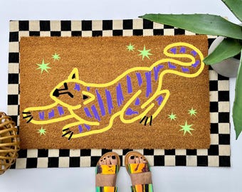 May include: A rectangular brown coir doormat featuring a stylized yellow cat illustration with purple stripes and black outlines. The design includes several small light green four-pointed stars scattered around the cat on the textured surface.