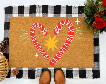 May include: A brown coir doormat with a black and white checkered border. The doormat has a red and white candy cane design in the shape of a heart with a yellow star in the center.