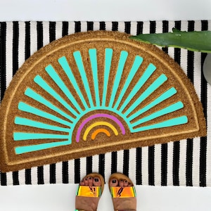 May include: A brown coir doormat with a turquoise sunburst design and a rainbow. The doormat is placed on a black and white striped rug. A woven basket and a potted plant are visible in the background.
