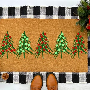 May include: A brown coir doormat with five green Christmas trees painted on it. The trees have red and green ornaments. The doormat is on a black and white checkered rug.