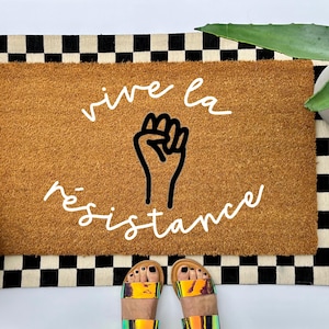 May include: A brown coir doormat with a black and white checkered border. The doormat has the text "vive la resistance" written in white cursive font with a black fist raised in the center.