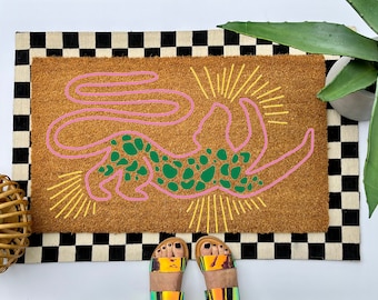 May include: A rectangular brown coir doormat featuring a stylized pink outline of a leaping leopard with green spots. The design includes yellow radiating sunburst lines above and below the animal, set against a black and white checkered border.