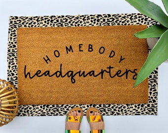 May include: A brown doormat with a black leopard print border. The doormat has the text "Homebody Headquarters" in black cursive lettering.