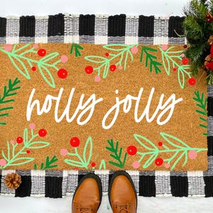 May include: A brown coir doormat with a black and white buffalo check border. The doormat has a green and red holly design and the text "holly jolly" written in white.