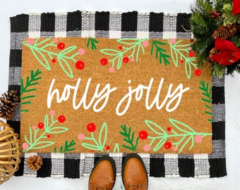 May include: A brown coir doormat with a black and white buffalo check border. The doormat has a green and red holly design and the text "holly jolly" written in white.