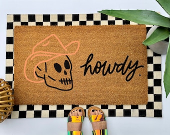 May include: A rectangular doormat with a brown coir center and a black and white checkered border. The mat features a skull wearing a cowboy hat and the word "howdy" in black script. The mat is placed on a white surface.