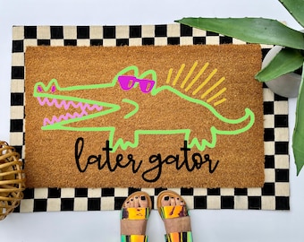 May include: A rectangular brown coir doormat featuring a neon green outline of an alligator wearing bright pink sunglasses. The alligator has a yellow spiky crest and pink teeth. The text "later gator" is printed in black cursive script.