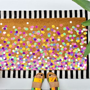 May include: A doormat with a brown background and a design of colorful confetti. The confetti is in various shades of pink, purple, yellow, green, and white.