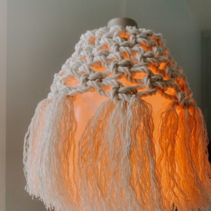 May include: An illuminated essential oil diffuser with a cream-colored macrame cover and fringe. The diffuser emits a warm, orange glow, creating a cozy ambiance. The base of the diffuser is a neutral color, with two buttons visible.