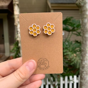 May include: Two yellow and white honeycomb-shaped stud earrings on a brown card. The earrings are held by a hand in front of a blurred background of a house and greenery. The card has a logo that says "The Green Porch".