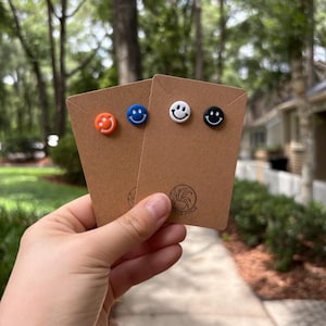 May include: Four pairs of smiley face stud earrings in orange, blue, white, and black. Each pair is mounted on a small, brown cardboard display card. The earrings are held in a hand against a blurred background of trees and a walkway.