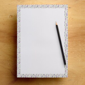 May include: A white notepad with a lavender floral border and a black pencil on a wooden surface. The notepad's border features a repeating pattern of purple and green floral elements. The pencil is positioned diagonally across the notepad.