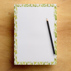 May include: A white notepad with a green leaf border and a black pencil on a wooden surface. The notepad has a watercolor design of green leaves and yellow accents. The pencil is positioned diagonally across the notepad.