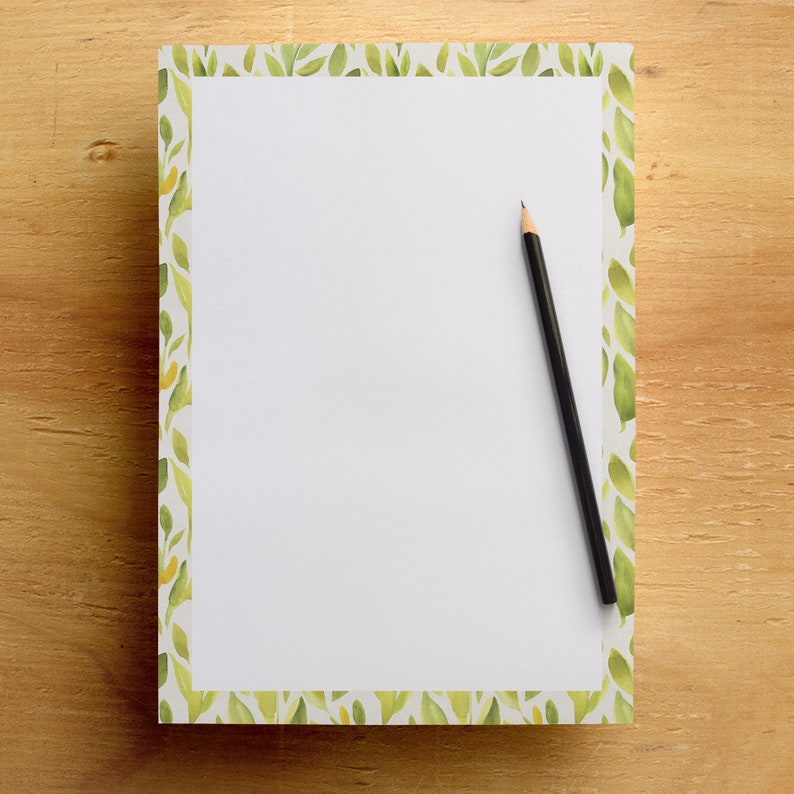 May include: A white notepad with a green leaf border and a black pencil on a wooden surface. The notepad has a watercolor design of green leaves and yellow accents. The pencil is positioned diagonally across the notepad.