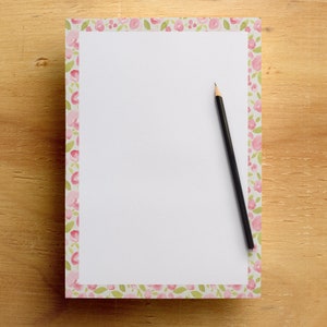 May include: A floral-bordered notepad with a blank white page and a black pencil. The notepad features a delicate pattern of pink flowers and green leaves. The pencil is positioned diagonally across the notepad, ready for writing.