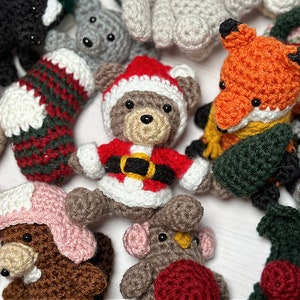 K&ouml;nnte beinhalten: Eine Sammlung handgefertigter geh&auml;kelter Weihnachtsornamente, darunter ein Strumpf, ein B&auml;r in einem Weihnachtsmannkost&uuml;m, ein Fuchs, ein Pilz und ein Rentier. Die Ornamente sind aus verschiedenen Garnfarben gefertigt, darunter Rot, Gr&uuml;n, Wei&szlig;, Braun und Schwarz.