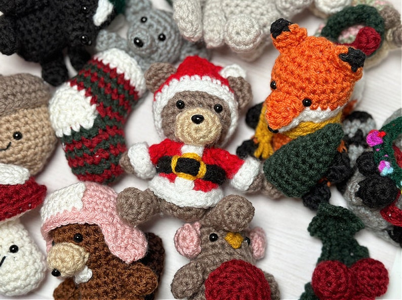 K&ouml;nnte beinhalten: Eine Sammlung handgefertigter geh&auml;kelter Weihnachtsornamente, darunter ein Strumpf, ein B&auml;r in einem Weihnachtsmannkost&uuml;m, ein Fuchs, ein Pilz und ein Rentier. Die Ornamente sind aus verschiedenen Garnfarben gefertigt, darunter Rot, Gr&uuml;n, Wei&szlig;, Braun und Schwarz.