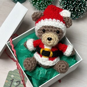 K&ouml;nnte beinhalten: Ein kleiner geh&auml;kelter brauner B&auml;r, der einen rot-wei&szlig;en Weihnachtsmann-Anzug mit einem schwarzen und gelben G&uuml;rtel tr&auml;gt. Der B&auml;r sitzt in einer wei&szlig;en Geschenkbox mit gr&uuml;nem Seidenpapier.