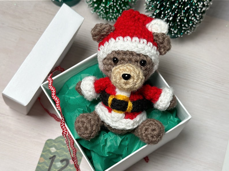 K&ouml;nnte beinhalten: Ein kleiner geh&auml;kelter brauner B&auml;r, der einen rot-wei&szlig;en Weihnachtsmann-Anzug mit einem schwarzen und gelben G&uuml;rtel tr&auml;gt. Der B&auml;r sitzt in einer wei&szlig;en Geschenkbox mit gr&uuml;nem Seidenpapier.