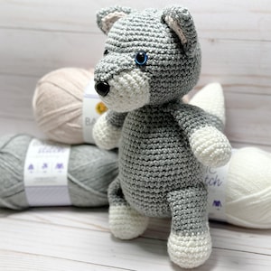 May include: A gray crocheted wolf plush toy with blue eyes and a white crocheted tail. The toy is sitting on a white wooden surface with three balls of yarn in the background.